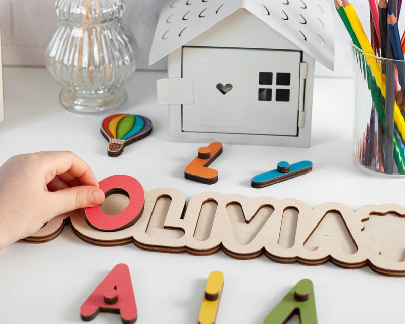May include: Wooden name puzzle featuring the name Olivia in large letters. The puzzle pieces are colourful and have a variety of shapes. The puzzle is on a white surface with other wooden toys.