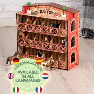 Puede incluir: Un juguete de establo de caballos de madera roja y marrón con múltiples puestos, con figuras de caballos en miniatura. El establo tiene el texto "Bryce's Stable" y está decorado con pequeños árboles. La imagen también incluye el texto "Personalización disponible en todos los idiomas."