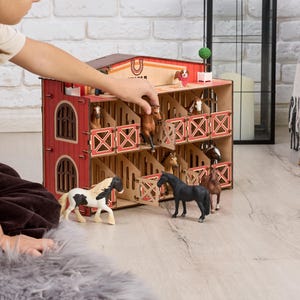 Puede incluir: Un establo de juguete de madera de dos pisos con paneles rojos y de madera natural, que cuenta con varios establos con puertas batientes. La estructura incluye varias figuras de caballos en miniatura de varios colores, incluidos negro, blanco y marrón, colocados dentro y fuera.