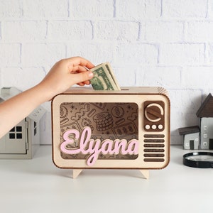 A wooden retro TV-shaped piggy bank with a slot on top for coins. The front of the bank has a clear plastic window with a black and white design of money and the name "Elyana" written in pink.