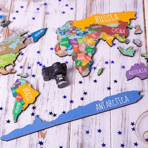 May include: A colorful wooden world map puzzle with names of countries like Russia, China, Brazil, and Australia. The puzzle pieces are arranged on a white wooden surface with blue star-shaped confetti scattered around. A small black camera is also on the surface.