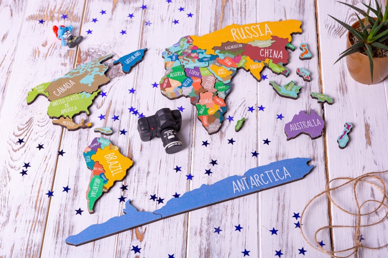 May include: A colorful wooden world map puzzle with names of countries like Russia, China, Brazil, and Australia. The puzzle pieces are arranged on a white wooden surface with blue star-shaped confetti scattered around. A small black camera is also on the surface.