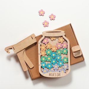 Può includere: Un salvadanaio in legno a forma di barattolo con gettoni a forma di fiore colorati. Il barattolo ha una fessura nella parte superiore ed è etichettato "MILA'S JAR" in basso. Il barattolo è parzialmente riempito con gettoni a forma di fiore rosa, arancione, verde, blu e viola.