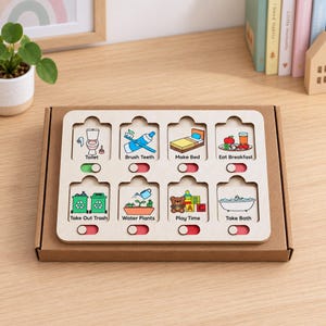 Puede incluir: Una tabla de tareas de madera clara con ocho ranuras rectangulares que incluyen ilustraciones coloridas y etiquetas de texto como Toilet, Brush Teeth, Make Bed, Eat Breakfast, Take Out Trash, Water Plants, Play Time y Take Bath.