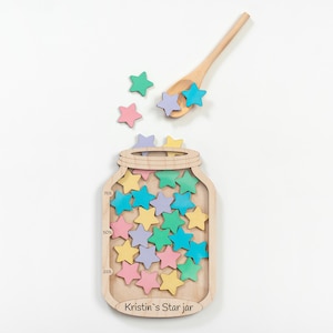 Può includere: Una tavola in legno a forma di barattolo con l'etichetta Kristin's Star jar e tacche per 25%, 50% e 75%, riempita con piccole stelle in legno color pastello. Un cucchiaio di legno abbinato tiene altre stelle sopra il barattolo.