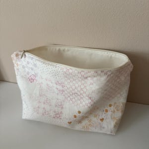 May include: A fabric pouch with a zipper closure featuring a cream-colored base and a repeating pattern of small, delicate floral and geometric motifs in shades of muted pink, soft yellow, and light gray, with a plain cream-colored interior lining.