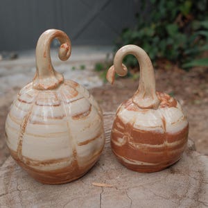 May include: Two ceramic pumpkins with a glossy finish, featuring a marbled cream and terracotta brown color pattern. Each pumpkin has a distinct, curved, elongated stem extending from the top, with visible ridges along the rounded bodies.