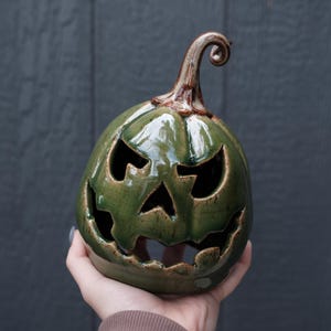 May include: A glossy olive green ceramic jack-o'-lantern with a carved face featuring triangular eyes, a triangular nose, and a jagged mouth. The pumpkin has a brown, curled stem at the top and a smooth, reflective finish throughout the surface.