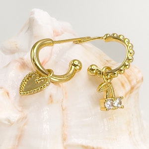 May include: A pair of gold hoop earrings with a heart charm and a cherry charm. The earrings are on a white seashell.