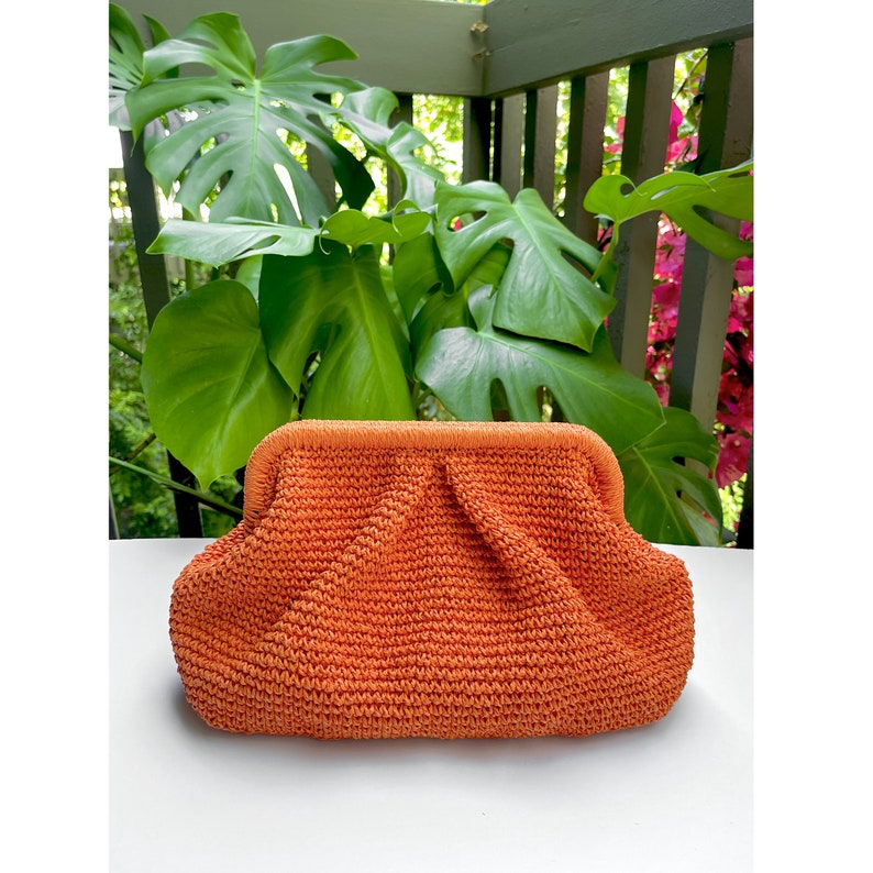 Puede incluir: Un bolso de mano tejido de color naranja brillante con un dise&ntilde;o fruncido. El bolso est&aacute; hecho de un material texturizado y tiene una forma redonda y lisa.