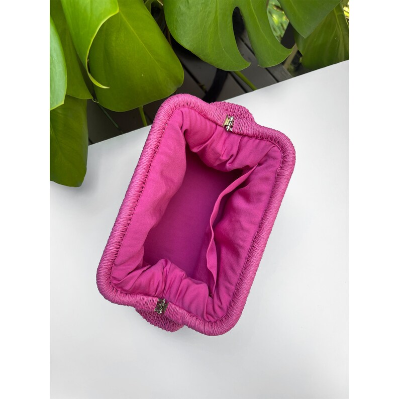 Puede incluir: Un bolso de mano tejido de color rosa con un forro de tela rosa. El bolso tiene una cremallera y una forma rectangular.
