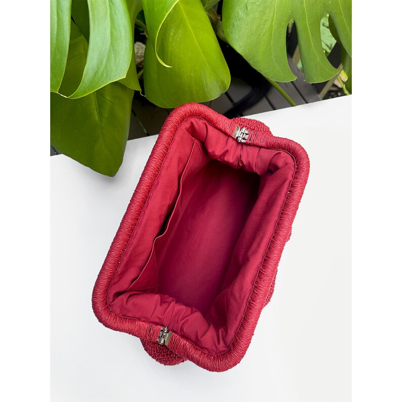 Puede incluir: Una cartera de mano tejida de color rojo con forro de tela y cierre de broche plateado.