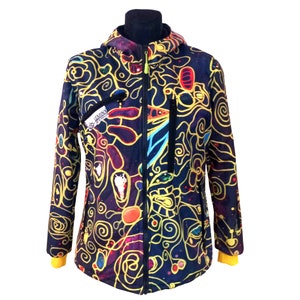 Op de afbeelding: Een zwarte hoodie met een kleurrijk abstract ontwerp met gele, rode, blauwe en groene lijnen en vormen. De jas heeft een ritssluiting en twee zakken.