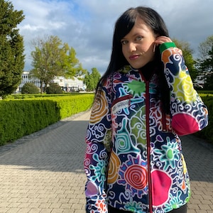 May include: A colourful, abstract patterned jacket with a zip closure. The jacket has a green hood and a red zip. The pattern includes swirls, circles, and other abstract shapes in various colours, including red, yellow, blue, green, and purple.