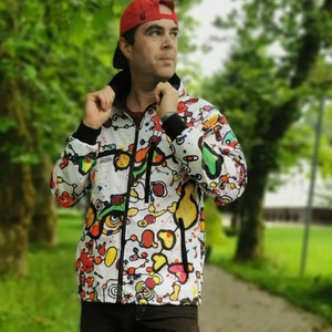 May include: A white jacket with a colorful abstract design. The jacket has a black zipper and a black hood. The person wearing the jacket is wearing a red baseball cap.
