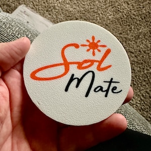 May include: A white round coaster with the words "Sol Mate" in black script. The word "Sol" is in orange with a small orange sun graphic above it.