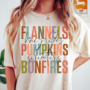 Può includere: Una t-shirt a maniche corte color avorio con una stampa grafica dall'aspetto vissuto che riporta le parole FLANNELS, hayrides, PUMPKINS, sweaters e BONFIRES in righe sovrapposte. Il testo utilizza una palette multicolore di toni arancio, verde e viola.