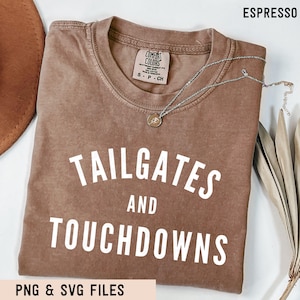May include: An espresso-coloured short-sleeve t-shirt with a crew neckline and a Comfort Colors brand label at the inner collar. The shirt features the white text "TAILGATES AND TOUCHDOWNS" printed in a bold, sans-serif font across the chest area.