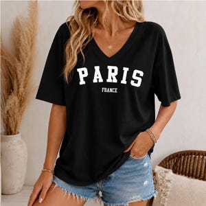 May include: A black short-sleeved t-shirt with a V-neckline featuring the white text PARIS FRANCE printed in a bold collegiate font across the chest. The shirt has a relaxed fit and is paired with light blue denim shorts with frayed hems.