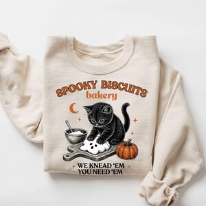 Pode incluir: Uma camisola de gola redonda cor de creme com um gráfico de um gatinho preto a amassar massa numa tábua de madeira. O texto diz SPOOKY BISCUITS bakery e WE KNEAD 'EM YOU NEED 'EM. Uma pequena taça, uma abóbora e uma lua crescente também estão representadas.