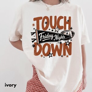 Könnte beinhalten: Ein elfenbeinfarbenes T-Shirt mit einem Grafikdruck, der den Text TOUCH DOWN in großen braunen Buchstaben zeigt. Zwischen den Wörtern befindet sich ein schwarzer Wimpel mit dem Text Friday Nights und einem Football-Motiv, umgeben von Sternen.