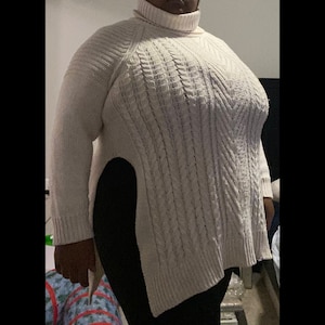 May include: A white cable knit sweater with a turtleneck and side slits. The sweater is made of a soft, comfortable material and has a relaxed fit.
