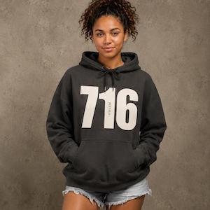 May include: A charcoal gray hooded sweatshirt featuring a large white graphic print of the number 716 with the word BUFFALO printed vertically inside the number one. The hoodie includes a front kangaroo pocket and matching black drawstring cords.