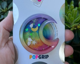 May include: A colorful PopGrip phone accessory with a rainbow and moon design. The PopGrip is made of clear resin with glitter and stars. The packaging says "PopGrip" and "Pull Twice to Grip".
