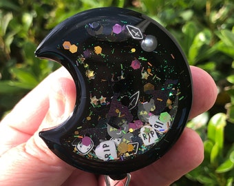 Puede incluir: Portagafete negro en forma de media luna con purpurina y pequeños amuletos. Los amuletos incluyen estrellas, corazones y la palabra "RIP".