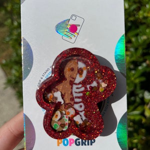 May include: A red glitter PopSocket with a gingerbread man design. The gingerbread man is half-skeleton and half-gingerbread. The PopSocket is on a white and holographic backing with the word "POPSOCKETS" at the top and "POPGRIP" at the bottom.