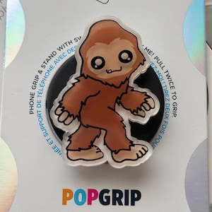 May include: A PopGrip phone grip featuring a cartoon Bigfoot character in shades of brown and tan. The grip is attached to a black base and is on a white background with the PopGrip logo in orange and blue.