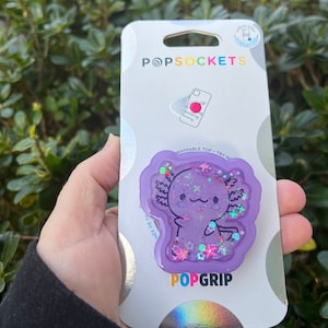 May include: Purple PopGrip phone grip with a cute axolotl design. The axolotl is surrounded by glitter and stars. The PopGrip is packaged on a white card with the PopSockets logo.
