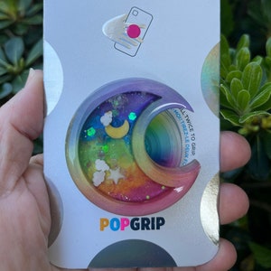 May include: A colorful PopGrip phone accessory with a rainbow and moon design. The PopGrip is made of clear resin with glitter and stars. The packaging says "PopGrip" and "Pull Twice to Grip".