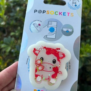 May include: A PopSockets PopGrip featuring a strawberry cow design. The cow is white with red accents and strawberries. The PopGrip is on a white and blue card with the PopSockets and PopGrip logos. The card also has instructions.