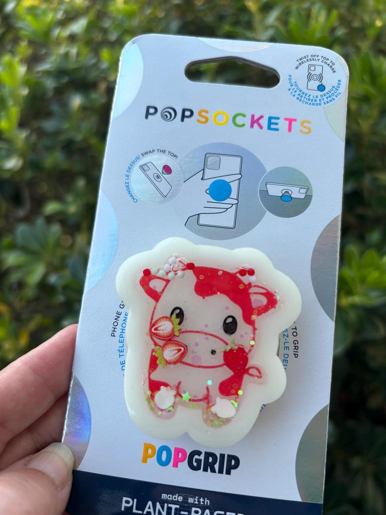 May include: A PopSockets PopGrip featuring a strawberry cow design. The cow is white with red accents and strawberries. The PopGrip is on a white and blue card with the PopSockets and PopGrip logos. The card also has instructions.