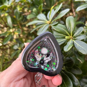 May include: Black heart-shaped resin charm with a black and white image of two hands reaching up to a moon with stars and a web design. The charm is attached to a clear plastic retractable badge holder.