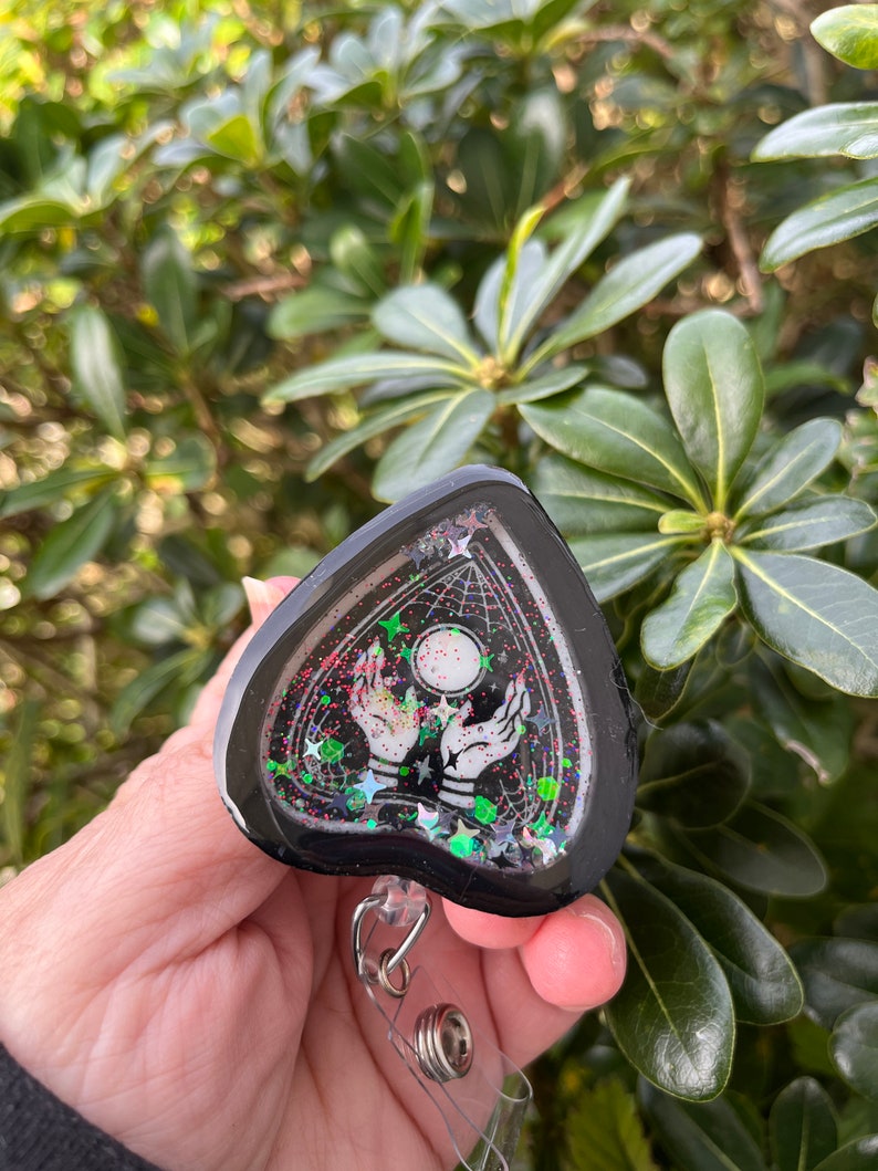 May include: Black heart-shaped resin charm with a black and white image of two hands reaching up to a moon with stars and a web design. The charm is attached to a clear plastic retractable badge holder.