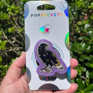 May include: A PopSockets PopGrip featuring a black raven design with gold stars and crescent moons on a purple background. The PopGrip is on a white and silver backing with the PopSockets logo.