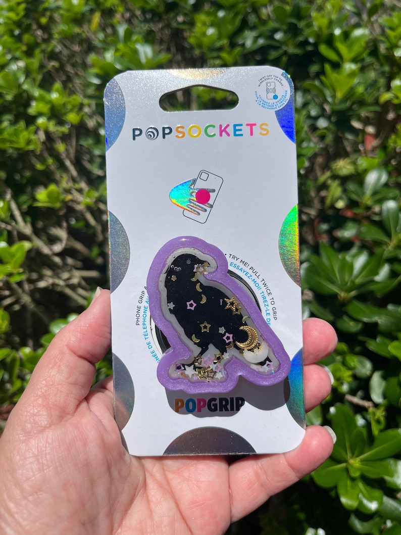May include: A PopSockets PopGrip featuring a black raven design with gold stars and crescent moons on a purple background. The PopGrip is on a white and silver backing with the PopSockets logo.