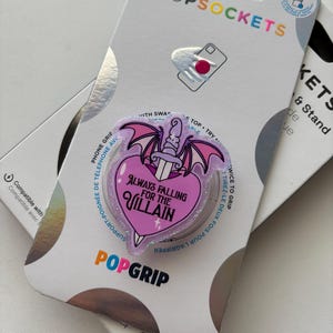 May include: A glittery, heart-shaped phone grip featuring a purple and pink design with a dagger piercing the heart and bat wings. The text reads Always Falling For The Villain. It is mounted on a white PopSockets brand retail display card.
