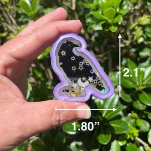 May include: A purple raven-shaped shaker charm, 2.1 inches tall and 1.80 inches wide, filled with a black background, gold and silver stars, and crescent moons. The charm is held in a hand against a backdrop of green foliage.