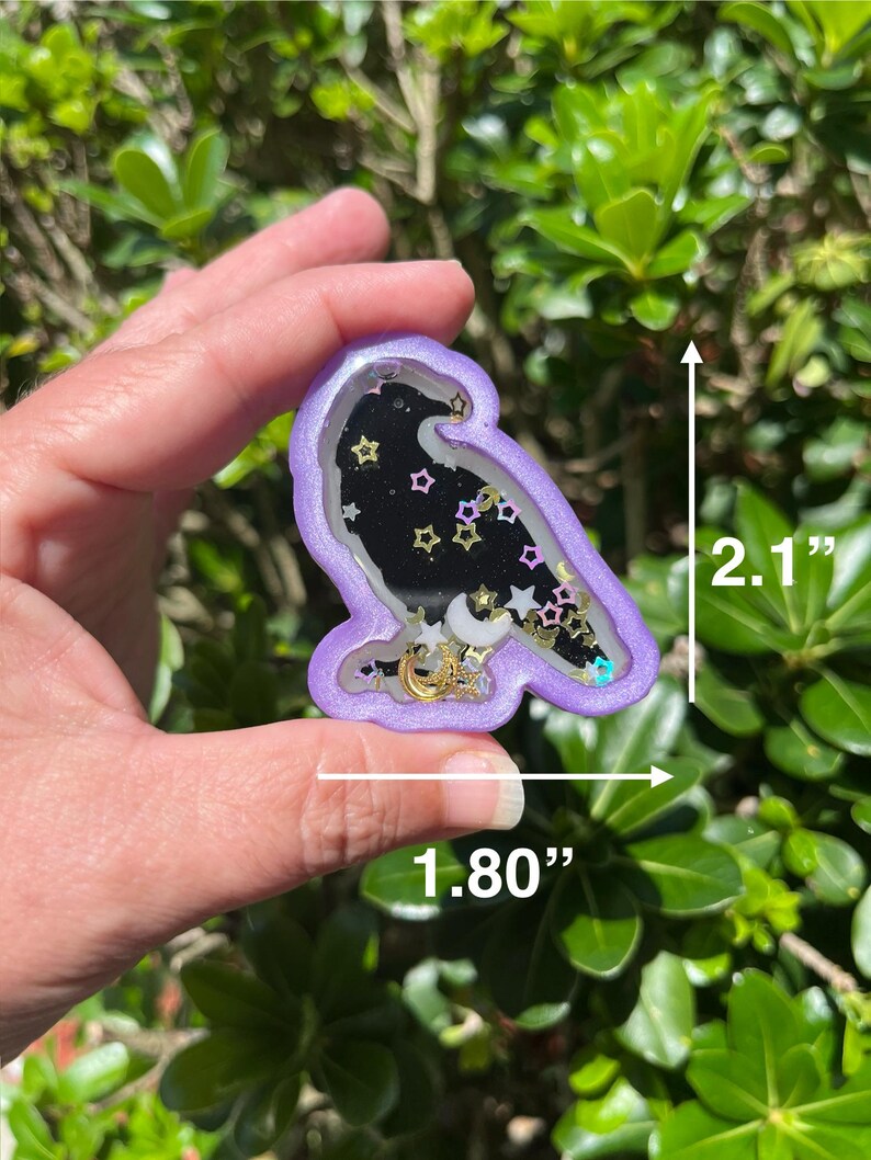 May include: A purple raven-shaped shaker charm, 2.1 inches tall and 1.80 inches wide, filled with a black background, gold and silver stars, and crescent moons. The charm is held in a hand against a backdrop of green foliage.