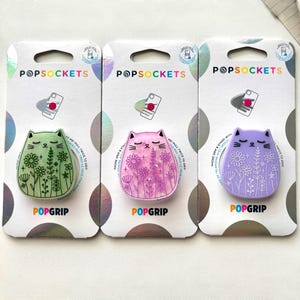 May include: Three PopSockets phone grips in green, pink, and purple, each shaped like a cat with floral designs. The PopSockets are on white and silver packaging with the brand name and product information.