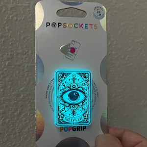 May include: A blue PopGrip phone grip with a black and white design of an eye with a star and a crescent moon. The text "The Evil Eye" is printed on the grip.