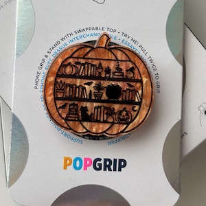 May include: A pumpkin-shaped phone grip with a swappable top featuring a black line art design of bookshelves filled with books, plants, a cauldron, a cat, and a jack-o'-lantern. The background is a swirled orange and gold glitter resin material.