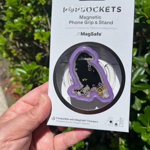May include: A PopSockets Magnetic Phone Grip & Stand in a package. The grip has a purple border with a black interior filled with gold stars and confetti. The packaging includes the text "MAGNETIC ADAPTER INCLUDED" and "Compatible with MagSafe Chargers."