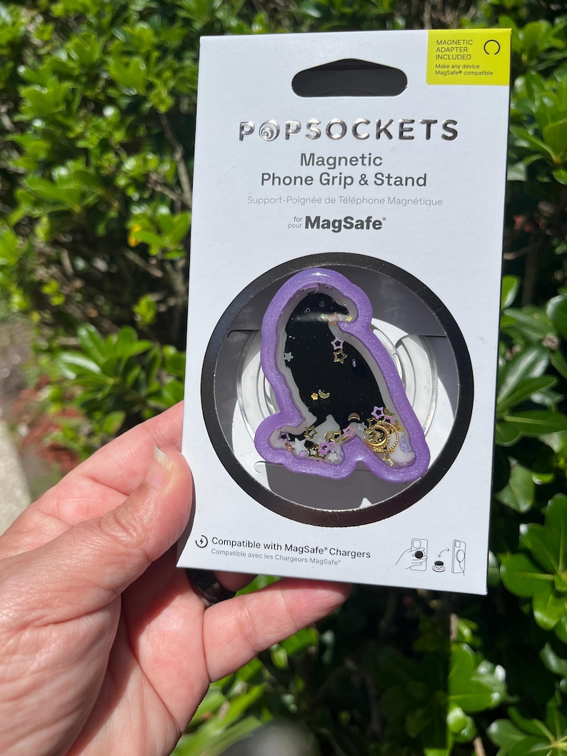 May include: A PopSockets Magnetic Phone Grip & Stand in a package. The grip has a purple border with a black interior filled with gold stars and confetti. The packaging includes the text "MAGNETIC ADAPTER INCLUDED" and "Compatible with MagSafe Chargers."