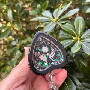 May include: Black and white ghost-shaped resin charm with glitter and a silver retractable badge reel. The charm features a hand reaching towards a glowing orb with stars in the background.