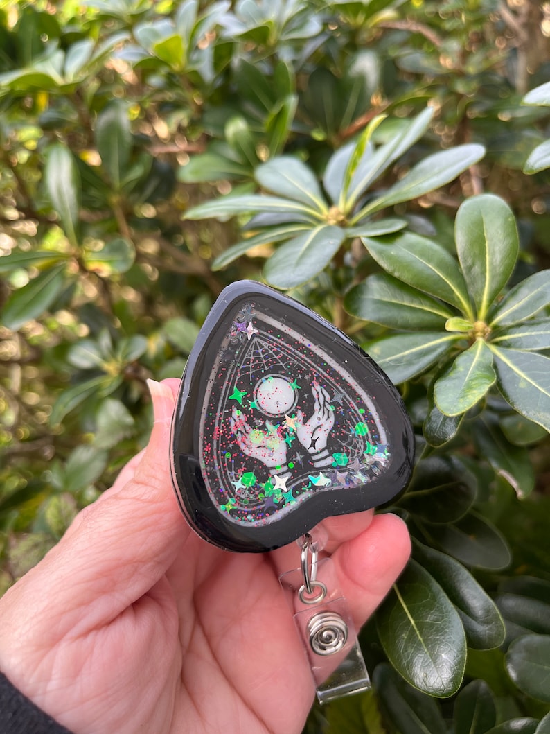 May include: Black and white ghost-shaped resin charm with glitter and a silver retractable badge reel. The charm features a hand reaching towards a glowing orb with stars in the background.