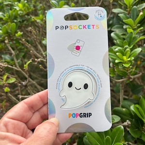 May include: A white PopSockets PopGrip with a ghost design. The PopGrip is on a white and iridescent backing with the PopSockets logo and the words "PopGrip" and "Phone Grip & Stand with Swappable Top".